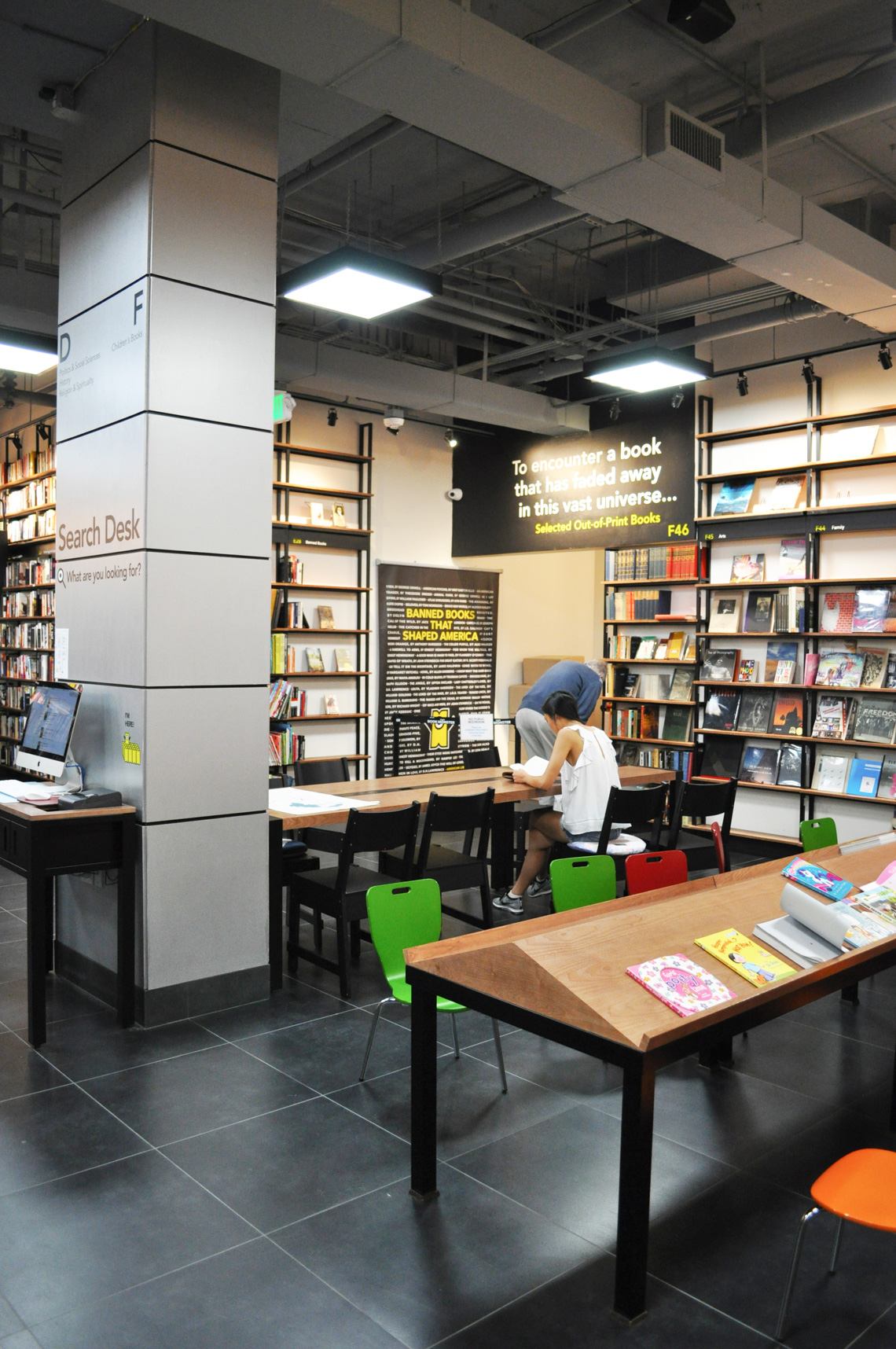 A building interior picture of book store. Book display wall, open ceiling with gray paint and dark gray tile floor finish. Wooden book display stands in the middle.
