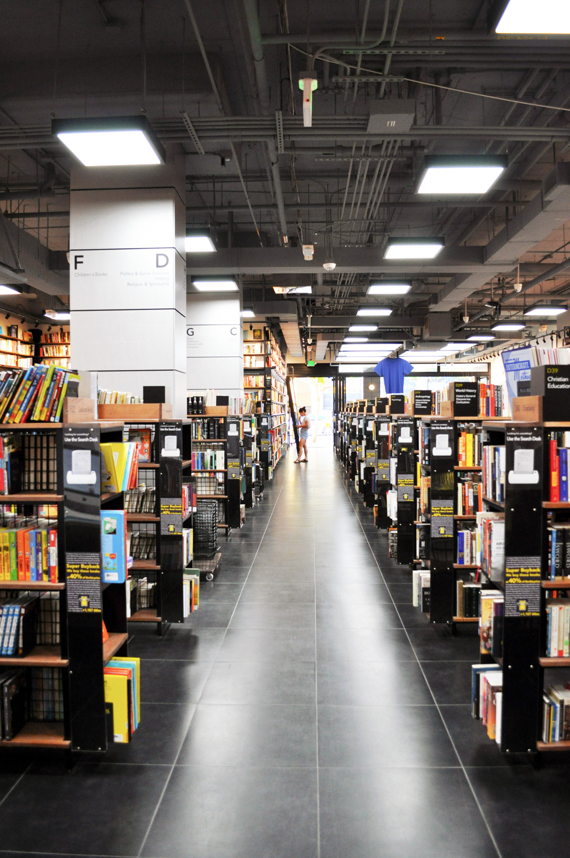 A building interior picture of book store. Book display wall, open ceiling with gray paint and dark gray tile floor finish. Wooden book display stands in the middle.