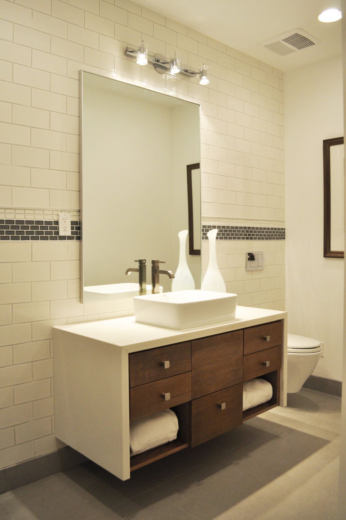 A building interior picture of the bathroom. gray tile floor and white tile wall. white vanity and mirror.