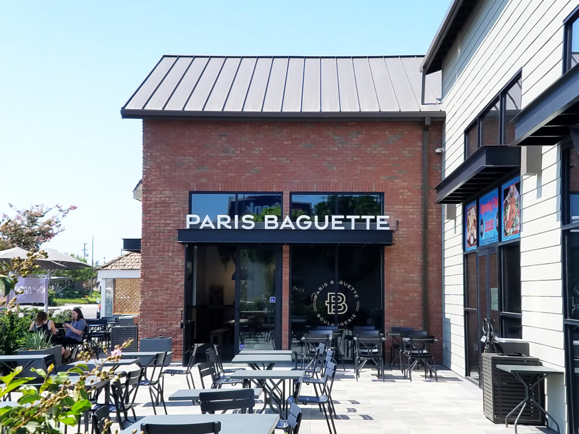 A building exterior picture of paris baguette stire with outdoor seating. Brick exterior wall and white exterior siding wall.