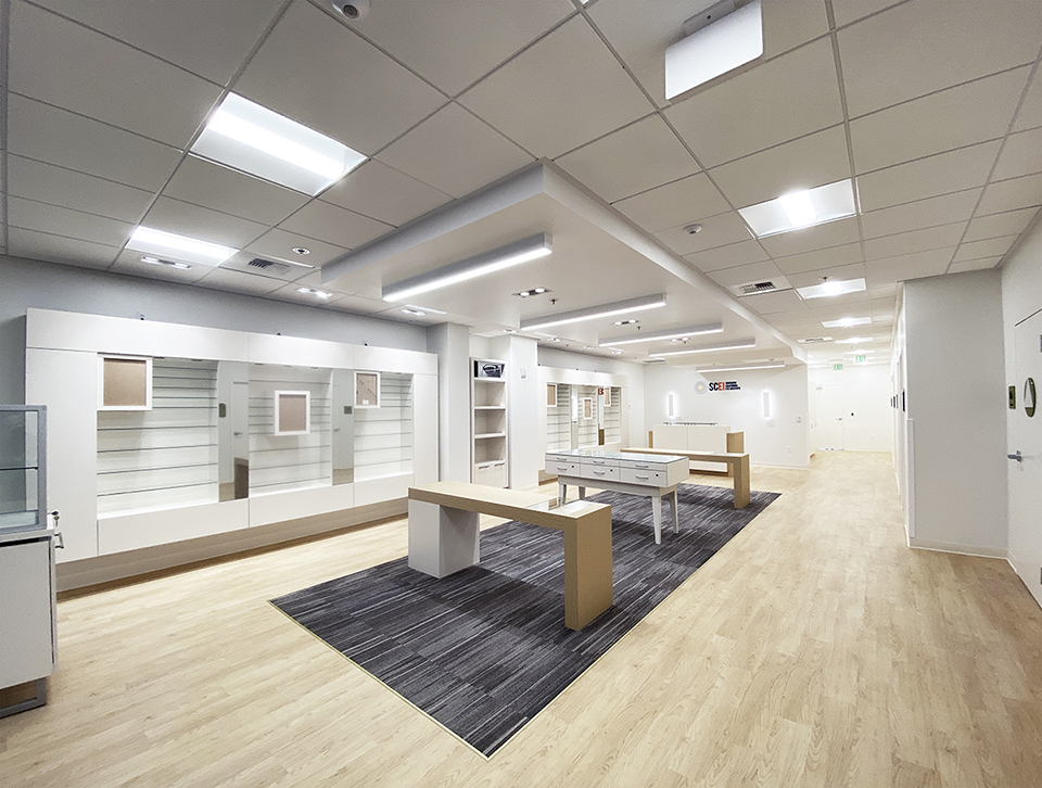 A building interior picture of optometry. White display wall, white ceiling and wood floor finish. Wooden display islands in the middle.