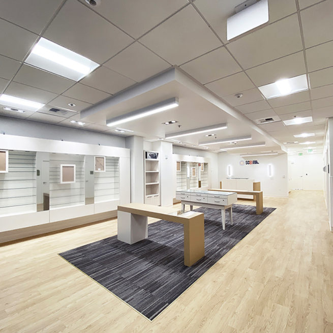 A building interior picture of optometry. White display wall, white ceiling and wood floor finish. Wooden display islands in the middle.